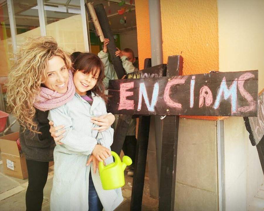 Dona abraçant una nena amb una regadora al costat d'un rètol que diu «Enciams»