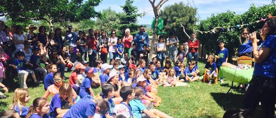 Grup nombrós d'infants asseguts a la gespa escoltant una monitora amb micròfon